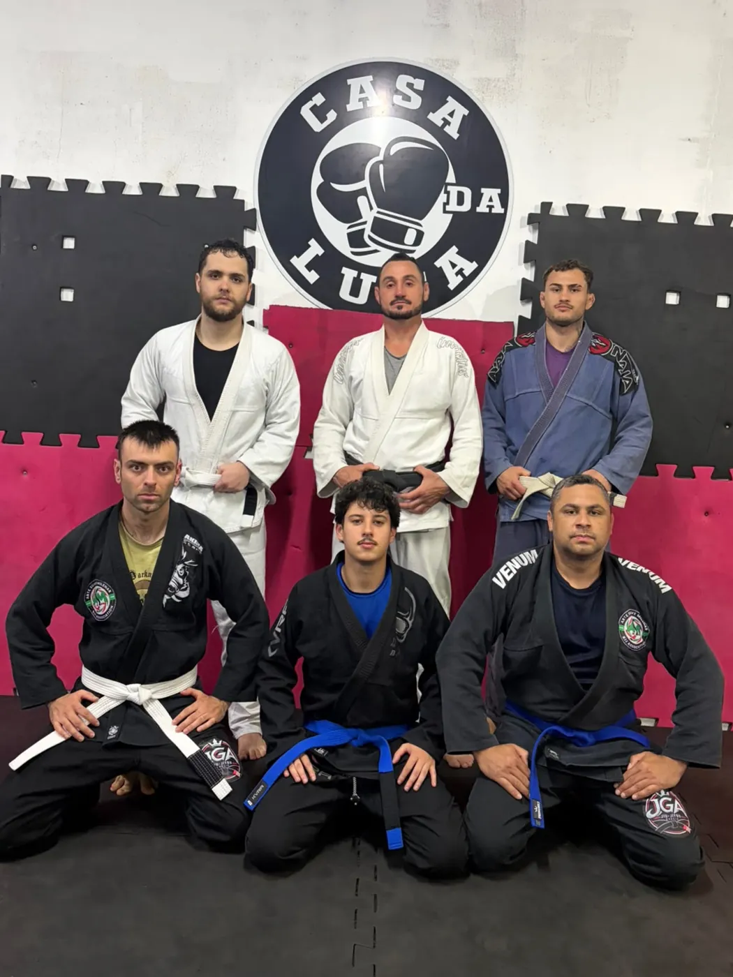 Professor e alunos de Jiu-Jítsu de quimono em frente à logo da Casa da Luta